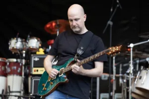 Gitarrist Alexander Stocker spielt seine grüne E-Gitarre, hinter ihm sind Schlagzeug und Percussion zu sehen.
