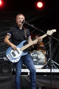 Bassist Jürgen Wernet singt am Mikrofon und spielt seinen Bass vor dem Schlagzeug mit dem Get-Wet-Logo.