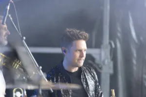 Trompeter Dennis Frommherz spielt konzentriert im glitzernden Bühnenoutfit, während Mikrofonständer und Instrumente den Vordergrund durchziehen.