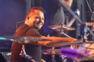 Schlagzeuger Andreas Scheerer greift lächelnd über sein Drumset, während violettes und blaues Bühnenlicht auf die Becken fällt.