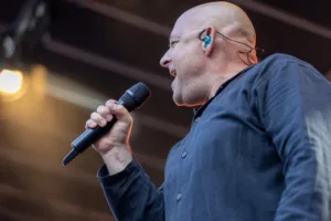 Sänger Sascha Spitz hält das Mikrofon nah ans Gesicht und hebt den freien Arm, während gelbe Lichtstrahlen hinter ihm auffächern.