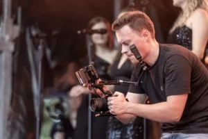 Der Kameramann richtet eine professionelle Filmkamera auf das Bühnengeschehen, während eine Sängerin unscharf im Hintergrund steht.