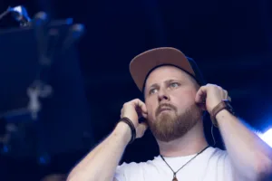 Posaunist Fabian Vogelbacher richtet mit beiden Händen seine In-Ear-Hörer, beleuchtet von kräftigem blauem Bühnenlicht.