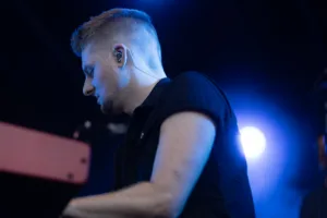 Keyboarder Tim Giron spielt an seinem Instrument, während ein heller blauer Scheinwerfer die dunkle Bühne durchbricht.