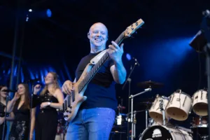 Bassist Steffen Vogt spielt lächelnd im Vordergrund, während Sängerinnen und Schlagzeug unscharf hinter ihm zu sehen sind.