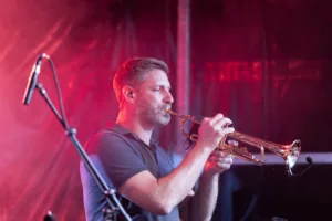 Trompeter Daniel Mertmann spielt vor rotem Bühnennebel, eingerahmt von Mikrofonständer und warm leuchtenden Scheinwerfern.