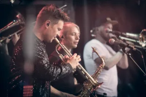 Trompeter Daniel Mertmann und weitere Musiker des Phil-It-Bläsersatzes spielen in einer Reihe, mit Saxofon und Mikrofonen im Hintergrund.