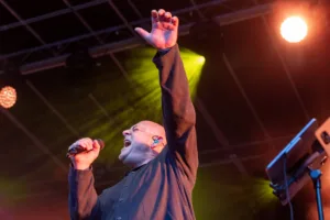 Sänger Sascha Spitz singt mit erhobenem Arm unter gelben Lichtstrahlen, die fächerförmig über die Bühne fallen.