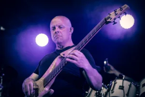 Bassist Steffen Vogt spielt seinen fünfsaitigen Bass vor dem Schlagzeug, von weißen und violetten Scheinwerfern beleuchtet.