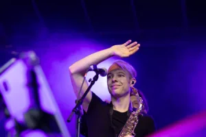 Saxofonist Max Marlier hebt eine Hand über die Augen und blickt vom Mikrofon aus ins Publikum unter violettem Licht.