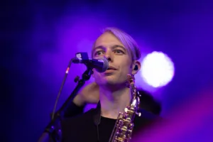 Saxofonist Max Marlier steht mit seinem Instrument am Mikrofon, während ein weiterer Musiker im violetten Bühnennebel wartet.