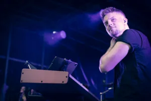 Keyboarder Tim Giron blickt vom Instrument zur Seite, während Tablet, Mikrofon und Bühnennebel den Arbeitsplatz einrahmen.