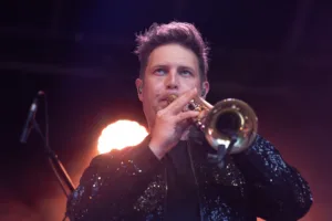 Trompeter Dennis Frommherz spielt frontal in die Kamera, während zwei orange Scheinwerfer hinter ihm aufleuchten.