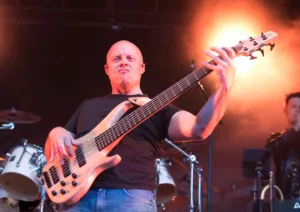 Bassist Steffen Vogt spielt am vorderen Bühnenrand, sein heller Bass hebt sich vor orangefarbenem Licht und dem Schlagzeug ab.