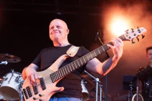 Bassist Steffen Vogt greift lächelnd in die Saiten seines hellen Basses, während ein orangefarbener Scheinwerfer hinter ihm leuchtet.