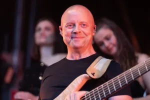 Bassist Steffen Vogt blickt lächelnd in die Kamera, seinen Bass vor der Brust und zwei Sängerinnen unscharf im Hintergrund.