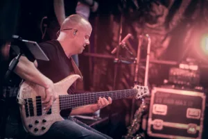 Bassist Steffen Vogt spielt im Sitzen am Bühnenrand, den langen Hals seines Basses quer durch das rötlich beleuchtete Bild geführt.