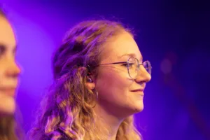 Sängerin Jana Maier blickt mit In-Ear-Hörern und Brille zur Bühne, von violettem Licht und einer unscharfen Sängerin eingerahmt.