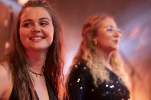 Sängerinnen Marleen Hoppe und Jana Maier stehen lächelnd nebeneinander, während eine dritte Sängerin unscharf hinter ihnen singt.
