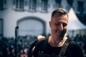 Gitarrist Sascha Happle steht mit geschlossenen Augen im warmen Abendlicht, während Publikum und historische Fassade hinter ihm verschwimmen.