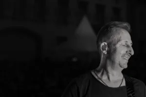 Gitarrist Sascha Happle lächelt im seitlichen Schwarzweißporträt, mit dunklem Bühnenhintergrund und dezentem Gegenlicht.