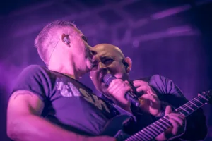 Gitarrist Sascha Happle lehnt sich zu Sänger Sascha Spitz, der unter violettem Licht dicht neben ihm ins Mikrofon singt.