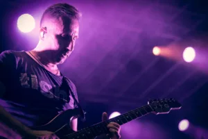 Gitarrist Sascha Happle spielt mit gesenktem Kopf ein Solo, während violetter Bühnennebel und Lichtpunkte den Hintergrund füllen.