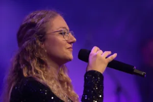 Sängerin Jana Maier singt mit Brille ins Handmikrofon, während violettes Licht ihr Profil vor dem dunklen Hintergrund hervorhebt.