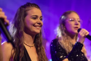 Sängerinnen Marleen Hoppe und Jana Maier singen gestaffelt unter violettem Licht, mit Mikrofonen und In-Ear-Hörern im Blick.