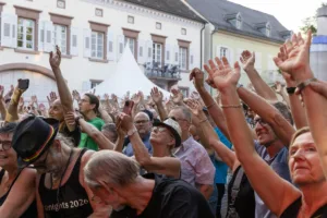 Das Publikum drängt sich mit erhobenen Händen vor der Open-Air-Bühne, während Musiker und Mikrofone am linken Rand zu sehen sind.