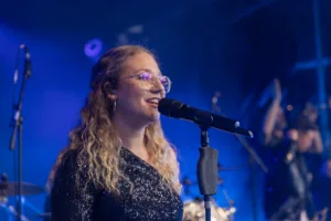 Sängerin Jana Maier singt mit Brille ins Bühnenmikrofon, während blaues Licht ihr Paillettenkleid und die Instrumente hinter ihr beleuchtet.