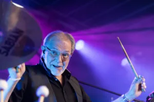 Percussionist Michael Scheerer hebt einen Drumstick über Becken und Trommeln, während violettes und blaues Licht sein Gesicht rahmt.