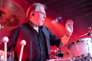 Percussionist Michael Scheerer hebt eine Hand über seine Trommeln, während rotes Bühnenlicht und weitere Musiker hinter ihm erscheinen.