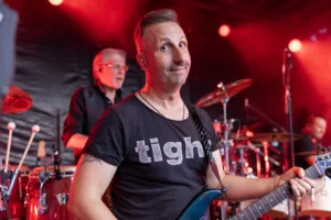 Gitarrist Sascha Happle blickt mit einem leichten Lächeln zur Seite, während Schlagzeug und rotes Bühnenlicht den Hintergrund füllen.