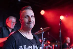Gitarrist Sascha Happle blickt nah an der Kamera vorbei, während rote Lichtpunkte und ein Musiker unscharf hinter ihm stehen.