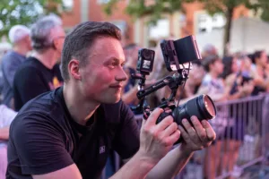 Der Kameramann kniet mit einer großen Filmkamera am Bühnenrand und richtet das Objektiv auf den Auftritt vor dem Publikum.