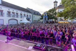 Das Publikum füllt den historischen Marktplatz bis zu den Fassaden, gesehen von der Bühne zwischen Monitoren und Mikrofonständern.