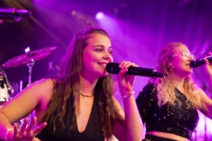 Sängerinnen Marleen Hoppe und Jana Maier singen an benachbarten Mikrofonen, eingerahmt von violettem Licht und Bühnennebel.
