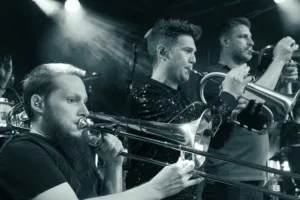 Drei Musiker des Phil-It-Bläsersatzes spielen Posaune, Saxofon und Trompete nebeneinander in einer kontrastreichen Schwarzweißaufnahme.