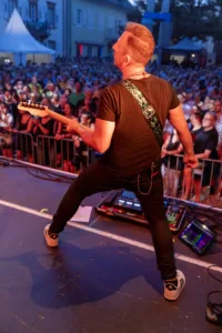 Gitarrist Sascha Happle spielt breitbeinig mit dem Rücken zur Kamera, während das Publikum den Platz vor der orange beleuchteten Bühne füllt.