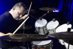 Schlagzeuger Andreas Scheerer spielt konzentriert hinter Becken und weißen Trommeln, den rechten Drumstick dicht am Kopf erhoben.