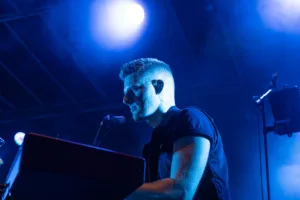 Keyboarder Tim Giron spielt im kühlen blauen Licht an seinem Instrument, während ein heller Scheinwerfer hinter seinem Kopf leuchtet.