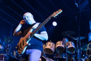 Bassist Steffen Vogt spielt seinen hellen Bass und berührt dabei seinen In-Ear-Hörer, mit Schlagzeug und blauem Licht im Hintergrund.
