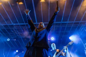 Sänger Sascha Spitz reckt beide Arme unter das Bühnendach, während Gitarrist und Bassist im blauen Licht hinter ihm spielen.
