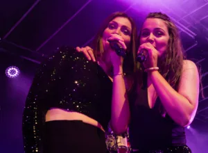 Sängerinnen Tamara Hoppe und Marleen Hoppe singen eng nebeneinander in ihre Mikrofone, die Arme im violetten Bühnenlicht umeinandergelegt.