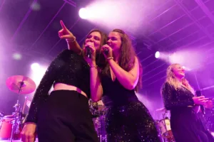 Sängerinnen Tamara Hoppe und Marleen Hoppe singen gemeinsam am Bühnenrand, von violettem Licht und einer weiteren Sängerin begleitet.